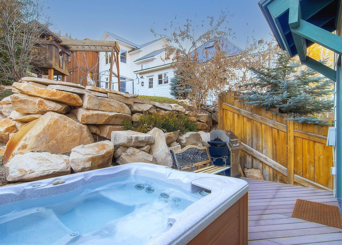 An outdoor hot tub and grill area with a wooden deck, surrounded by a rock wall and trees, with Woodside Manor visible in the background.