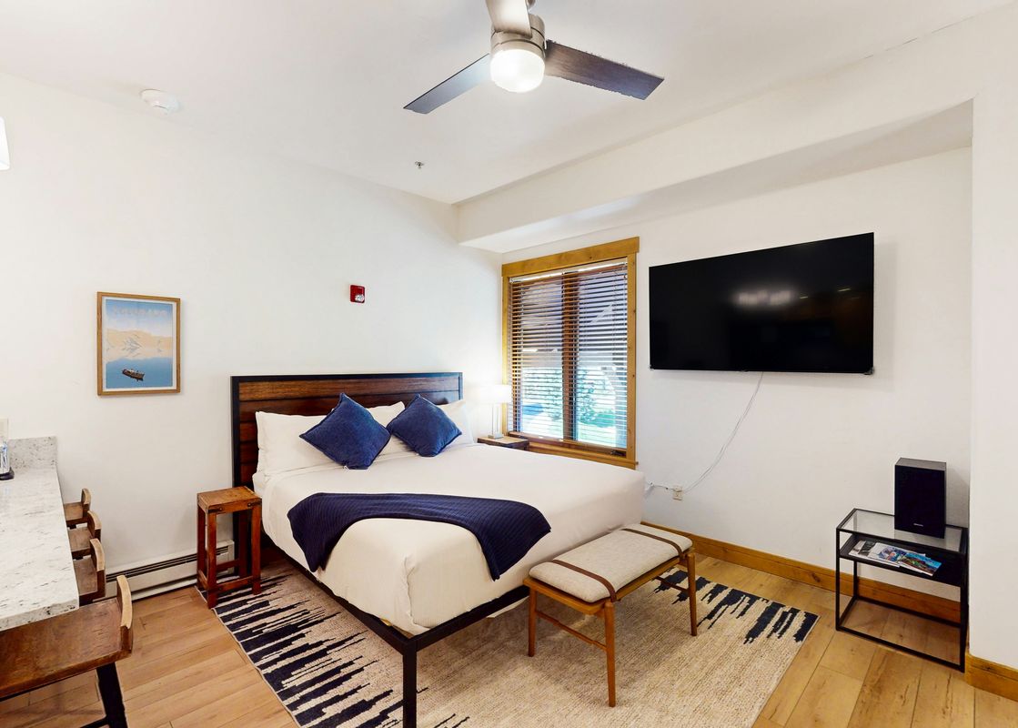 A cozy king-size bed with blue pillows and a navy throw blanket in EagleRidge Lodge 313's bedroom, featuring a flat-screen TV, a ceiling fan, and wooden blinds on the window.