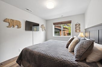 A serene bedroom at Pioneer Peak featuring a large bed with gray bedding, rustic wooden wall decor, and a window with a view of modern cabins.