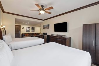 A hotel room with two queen beds, a dresser with a TV, a mini-fridge, microwave, and a dark wooden wardrobe. A ceiling fan is mounted above, and a separate living area with a sectional sofa and framed art is visible in the background.