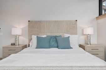 A cozy bedroom at One Riverfront Townhome 05 features a bed with a woven headboard, white and teal pillows, and matching silver table lamps on nightstands.