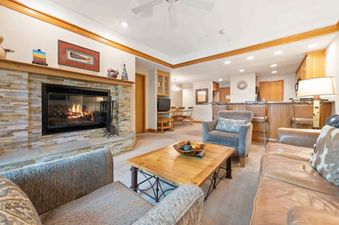 A cozy living room in The Charter G210 with a stone fireplace, a comfortable gray armchair, a brown leather couch, and a wooden coffee table.