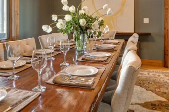 A rustic dining room table set with white plates and glasses, with a bouquet of white tulips in a vase in the center.