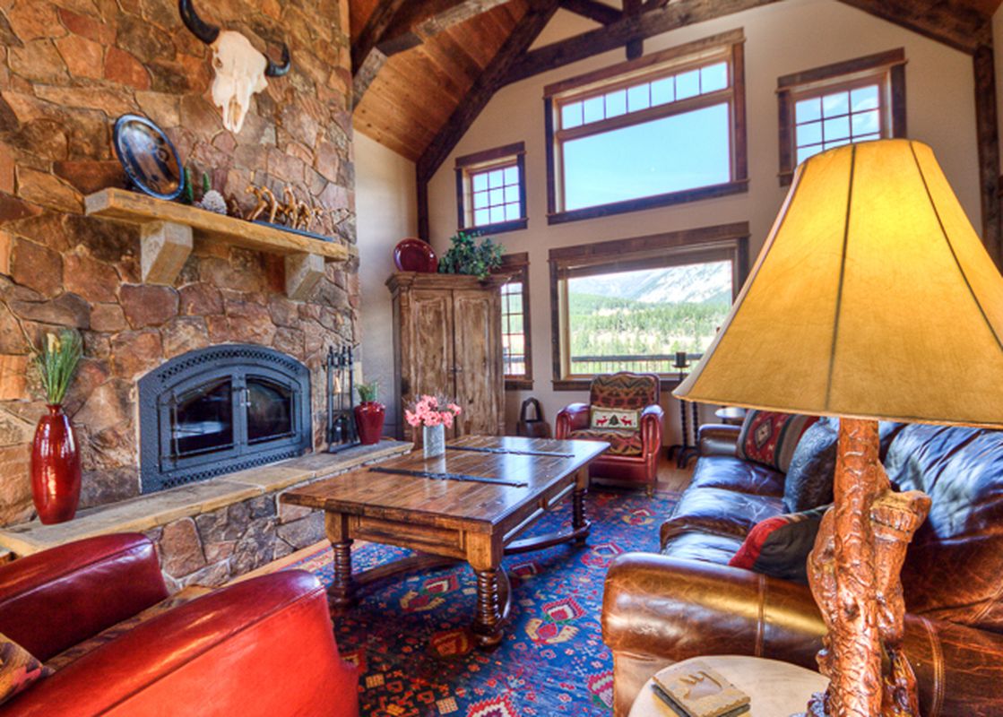 A cozy, rustic living room at Antler Ridge Lodge features a stone fireplace, leather furniture, and large windows with mountain views.