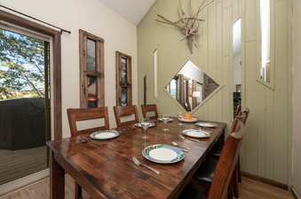 The dining area features a large wooden table with place settings, surrounded by chairs, and a sliding glass door leading to an outdoor deck.