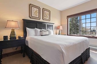 A king-sized bed with a black headboard and white linens sits in the bedroom of The Charter C1405, flanked by bedside tables and lamps, with a window offering an exterior view of trees and mountains.