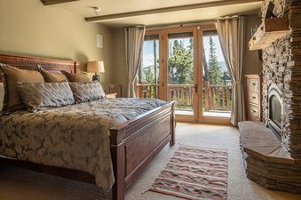 A luxurious bedroom featuring a wooden bed, a stone fireplace, and double doors opening to a balcony with mountain views.