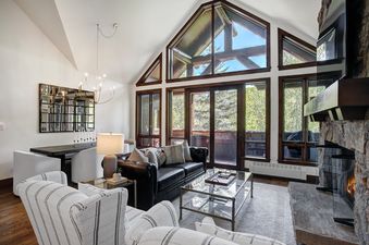 A spacious living area in Snow Cloud Lodge 715 features a black leather sofa and striped armchairs, a glass coffee table, a stone fireplace, and large windows with outdoor views. A dining area with a dark wood table and white chairs is also visible.