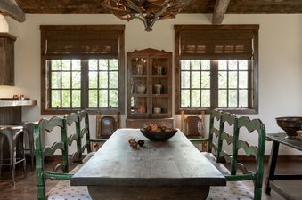 A rustic dining room features a large wooden table with green and white chairs, an antler chandelier, and wooden ceilings with exposed beams.
