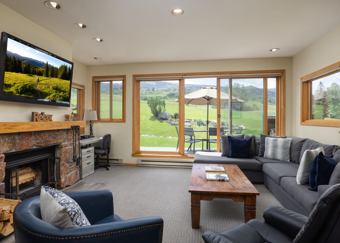 A spacious and inviting living room featuring a large sectional sofa, a coffee table, a fireplace, and a mounted TV, with large windows overlooking a scenic mountain landscape.