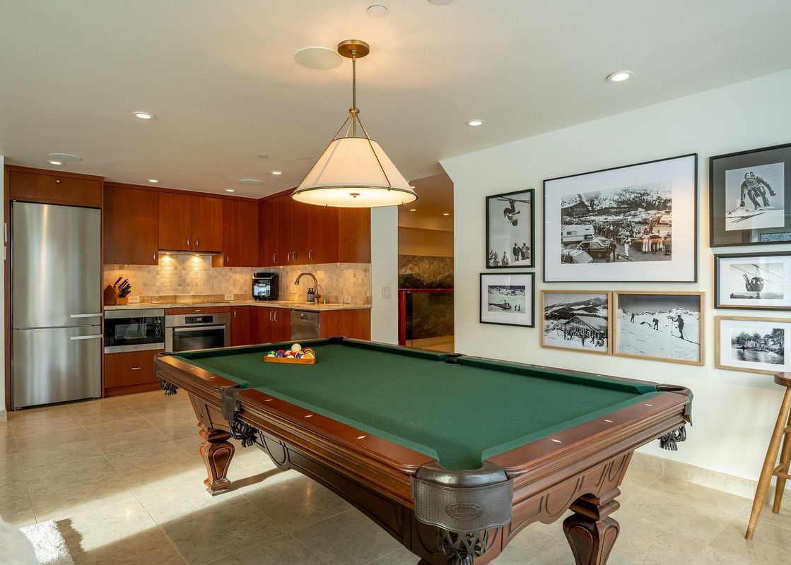 A large billiards table with green felt and wooden legs stands in a spacious room, with a modern kitchen in the background featuring stainless steel appliances and wooden cabinetry.