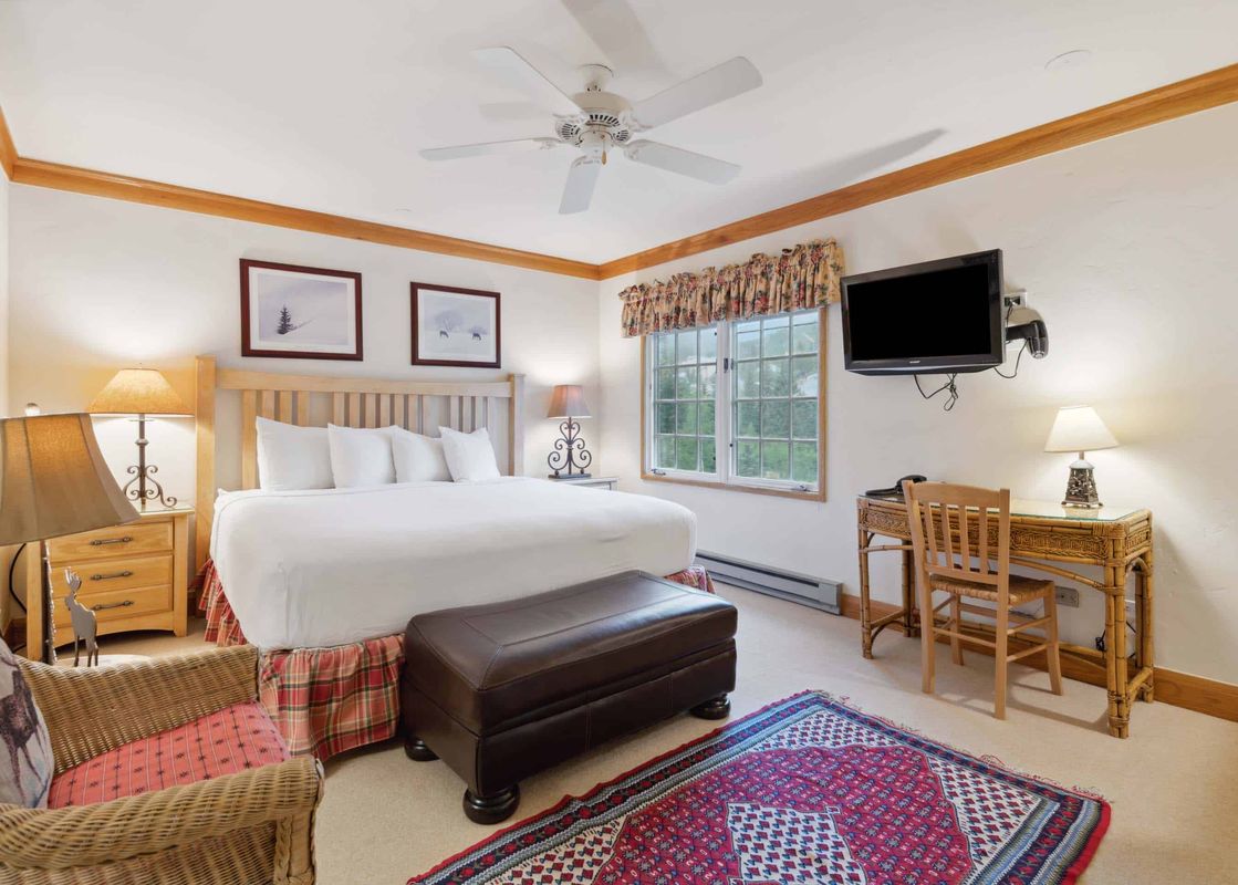 A master bedroom with a king-sized bed, two nightstands with lamps, a leather ottoman, a desk with a chair, and a mounted flat-screen TV.