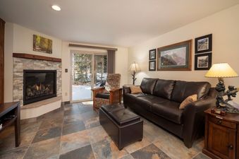 A cozy living room at Beaver Village 414 features a brown leather sofa, stone fireplace, and access to a snowy outdoor area.