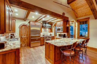 The kitchen at Bear Tracks Lodge features elegant wooden cabinetry, modern stainless steel appliances, a large granite island with seating, and rustic wooden beams.