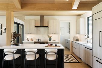 A modern kitchen with light wood cabinets, white countertops, and a large island with three bar stools, featuring a cowboy sculpture.