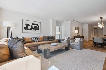 The living room of Solaris 6DE features a large sectional couch with throw pillows and a large coffee table with a  white bowl. The room is furnished with two armchairs with pillows and a wood coffee table. There is a painting on the wall and a large chan