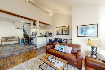 The spacious living room in Kutuk 310 features a comfortable leather sofa, a large coffee table, and a stylish lamp on a side table. The room is decorated with artwork on the walls and has a beautiful view of the surrounding mountains through the large wi