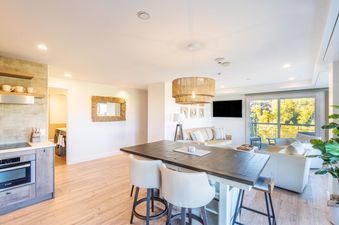 The Peaks Penthouse 434 kitchen and dining area features a large, dark wood dining table and bar chairs, with a modern, white and wooden bar counter on the side, and a fully equipped kitchen with stainless steel appliances, cupboards, and a microwave.