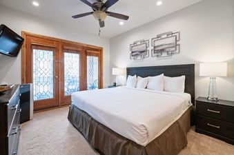 A cozy bedroom with a large bed, white linens, dark wood furniture, and artistic decorations, illuminated by soft lamps and natural light from decorative glass doors.
