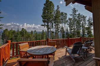 The private deck at Moonlight Mountain 1 Hidden Trail features a round dining table and chairs, with a backdrop of a pine forest and mountains under a blue sky.