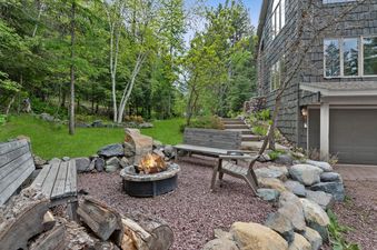 A cozy outdoor fire pit area at Elk Run with a burning fire, surrounded by rustic wooden benches and large rocks, set against a backdrop of lush green trees and a hillside.