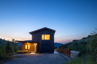 The Lodge Cabins Six Bedroom Mountain Cabin at dusk, showcasing its dark wood exterior, illuminated windows, and surrounding natural landscape with mountains and trees under a colorful sky.