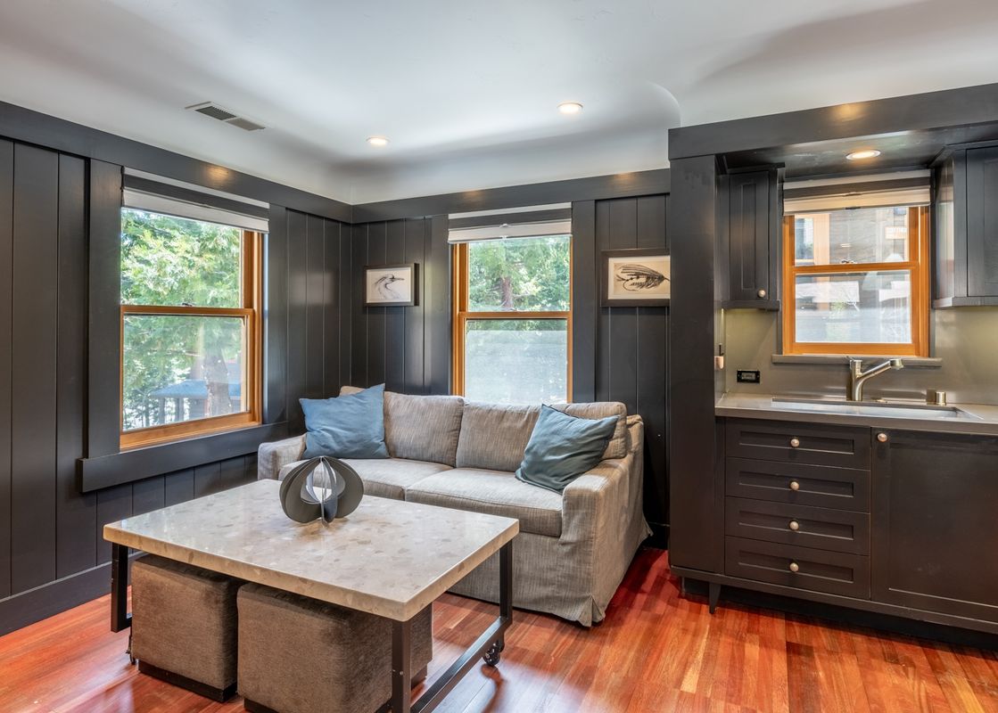 The living area of Cedar Crest Woodpecker Cottage 7 features dark wood paneling, a gray sofa with blue pillows, a coffee table with ottomans, and a compact kitchenette with a sink and dark cabinets.