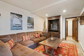 A cozy living room at The Charter E340 features a brown sectional sofa, a rustic wooden coffee table, a red patterned rug, two large framed winter art pieces, and a built-in entertainment center with a television.