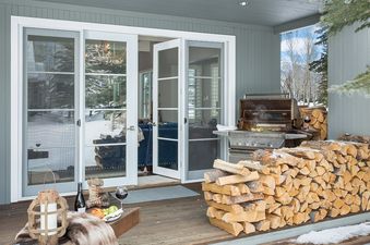A cozy outdoor patio with a BBQ grill, stacked firewood, and a bottle of wine with fruit on a wooden tray, set against a snowy landscape visible through glass doors.