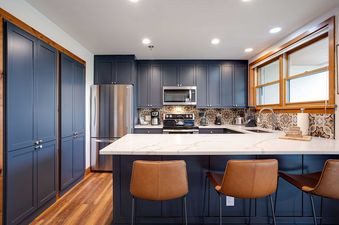 A modern kitchen at Chateau Chamonix CX211 featuring blue cabinetry, stainless steel appliances, white countertops, and a patterned backsplash, with two leather bar stools at the island and a window providing natural light.