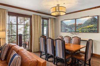 A cozy dining area at Elkhorn Premier 8405 with a large wooden table, striped and leather chairs, and a chandelier, offering a view of snow-covered mountains and ski lifts through glass doors.