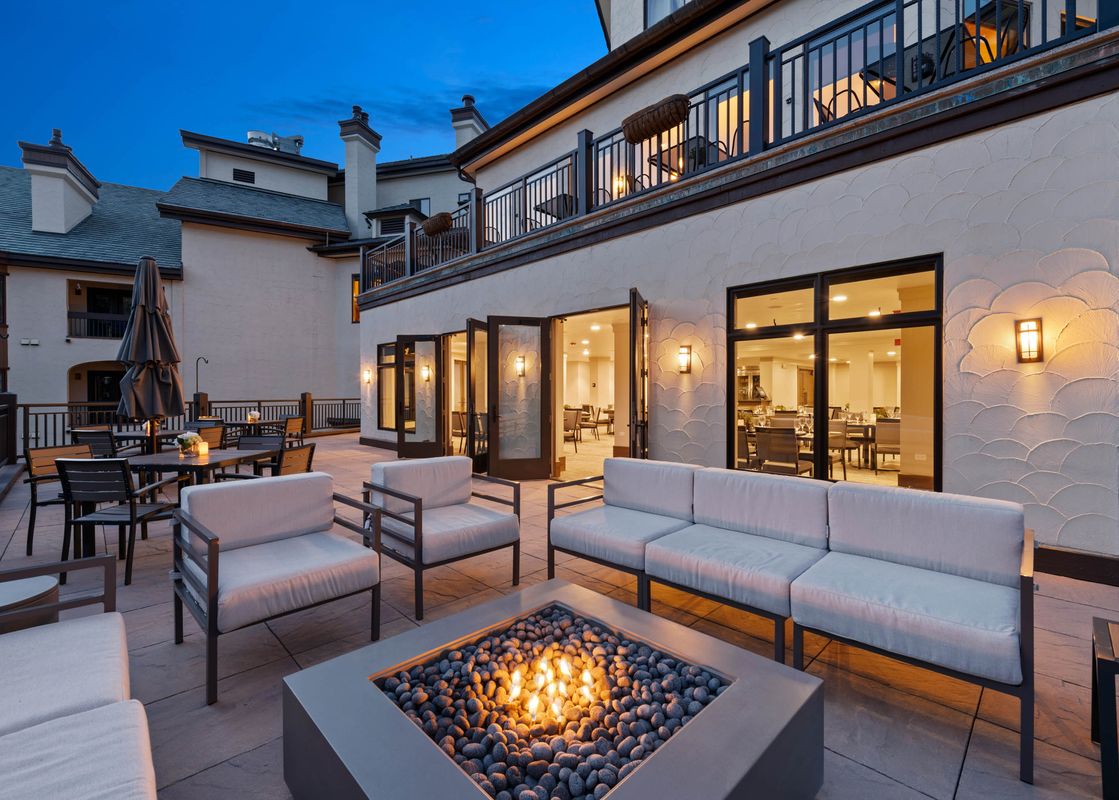 An evening view of The Charter C1105's outdoor patio area, featuring a cozy lounge arrangement with a lit fire pit, adjacent to an indoor dining space visible through open glass doors.