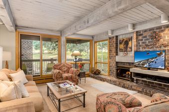 The inviting living room at Top of The Village Trails 208 features a plush sofa, a patterned armchair, a glass coffee table, and a brick fireplace with a television, all illuminated by natural light from large windows with outdoor views.