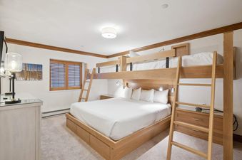 A bunk room at The Charter L032 features a queen bed, custom-built wooden bunk beds, reading lights, light wood furnishings, and a window with wooden blinds.