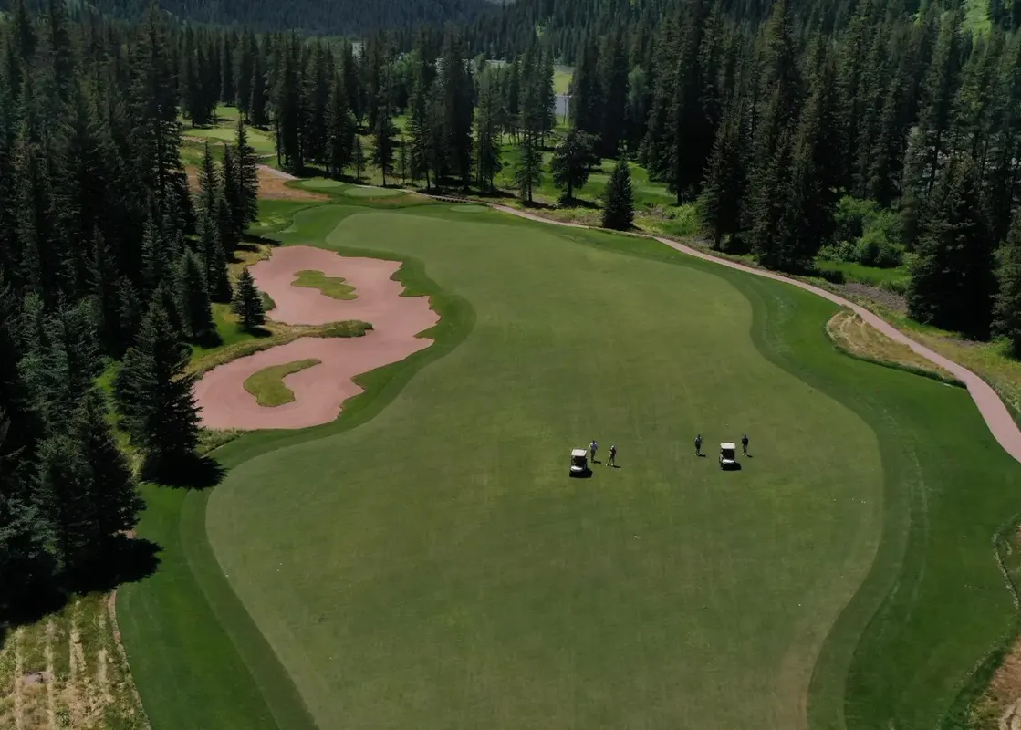 An aerial view of a vibrant green golf course, surrounded by a dense forest of evergreen trees, with several sand bunkers and a path winding through the landscape.