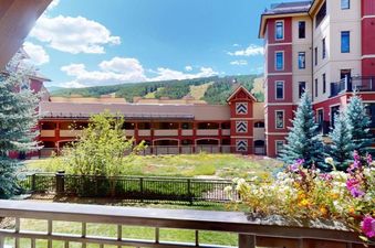 A vibrant summer view from the private balcony of Montaneros 217, featuring colorful flowers, resort buildings, and mountains under a blue sky.