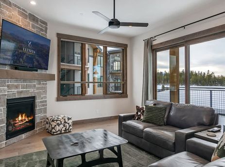 A cozy living room featuring a stone fireplace with a mounted flat-screen TV, a dark leather sofa, and large windows offering scenic outdoor views.