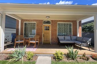 A front porch with a wooden floor, two wooden chairs and a couch and a wooden front door. The porch is covered by a wooden roof.
