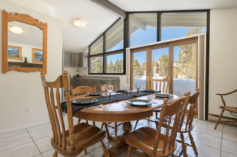 A large dining table with six wooden chairs sits in a brightly lit dining room, set with plates and glasses.