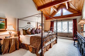 The bedroom at Bachelor Gulch Road 57W features a four-poster iron bed with patterned bedding, a vaulted wooden ceiling, and large windows overlooking a snowy landscape.