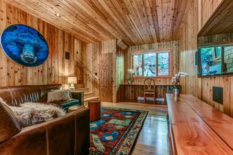 A cozy, wood-paneled living room featuring a large circular blue bear painting, a brown leather sofa, a vibrant patterned rug, and a built-in wooden desk with an outdoor view.