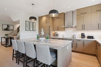 A modern kitchen in One Riverfront Townhome 07 features light wood cabinetry, stainless steel appliances, and a granite island with four bar stools and decorative plants.