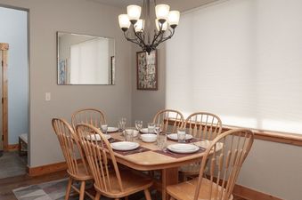 A bright dining area features a wooden table set with white dishes and clear glasses, surrounded by light wooden chairs, illuminated by an overhead chandelier.
