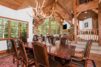 The dining area of Pine Meadow Estate features a long wooden table surrounded by brown leather chairs, a large antler chandelier, and large windows that look out onto the surrounding forest.