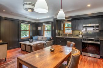 A modern open-concept living room and kitchen area with dark cabinetry, stainless steel appliances, a wooden dining table, and a comfortable sofa.