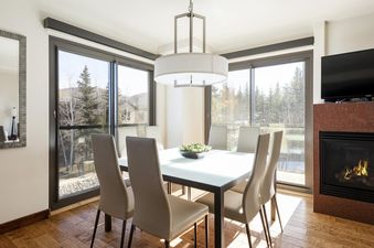 A dining room with a glass table, six chairs, and a large window with a view of a snow-covered forest.