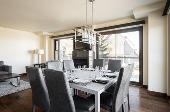 A modern dining area at Lowell 201B with a large table set for eight, a stylish chandelier, and floor-to-ceiling windows offering outdoor views.