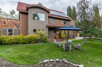 An inviting exterior of Parkway Treehouse Ketchum, showcasing its unique architecture, lush lawn, and a cozy patio setup with chairs and a blue umbrella, surrounded by mature trees.