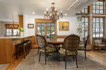 The dining area in Chalet 12 features a wooden table, plaid chairs, a chandelier, and large windows with an outdoor view.