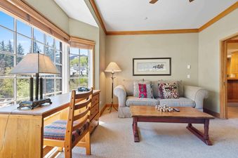 A cozy living room in The Charter E3305 with a large window overlooking a lush mountain landscape, a sofa with patterned pillows, a wooden coffee table, and a desk with a chair.